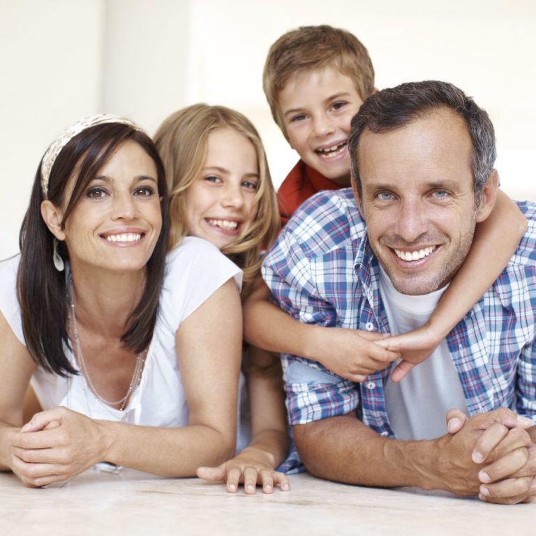 Eine Mutter und ein Vater, die sich mit ihren Armen auf einer hellen Oberfläche aufstützen. Der Vater wird von hinten von  seinem Sohn umarmt. Die Tochter lehnt sich bei der Mutter an.