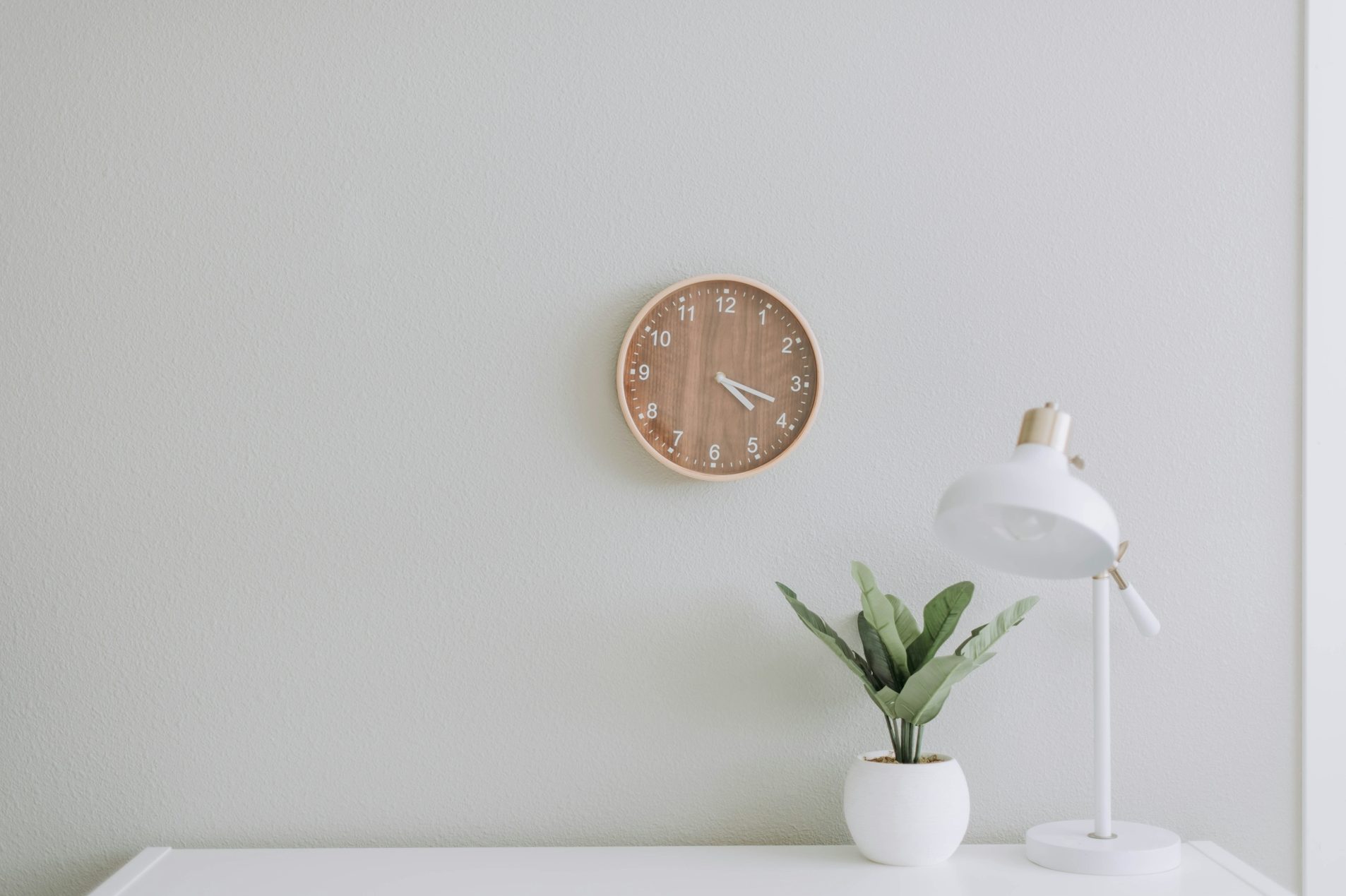 eine weiße Wand mit einer Uhr, einem Beistelltisch, einer Pflanze und einer Tischlampe